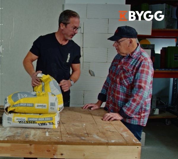 Två byggnadsarbetare vid arbetsbänk med gula murbruksäckar och K-Bygg-logga i verkstad