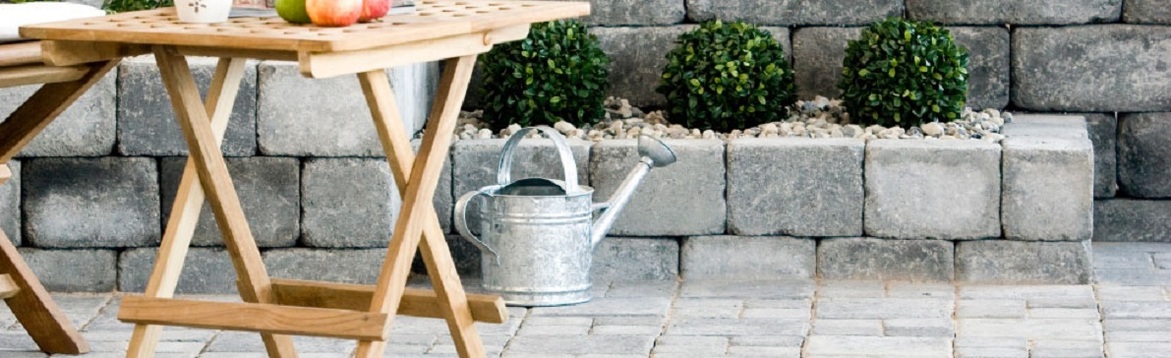 Utealtan med ljust träbord, röda äpplen, zinkvattenkanna framför grå stenmur och gröna buskar