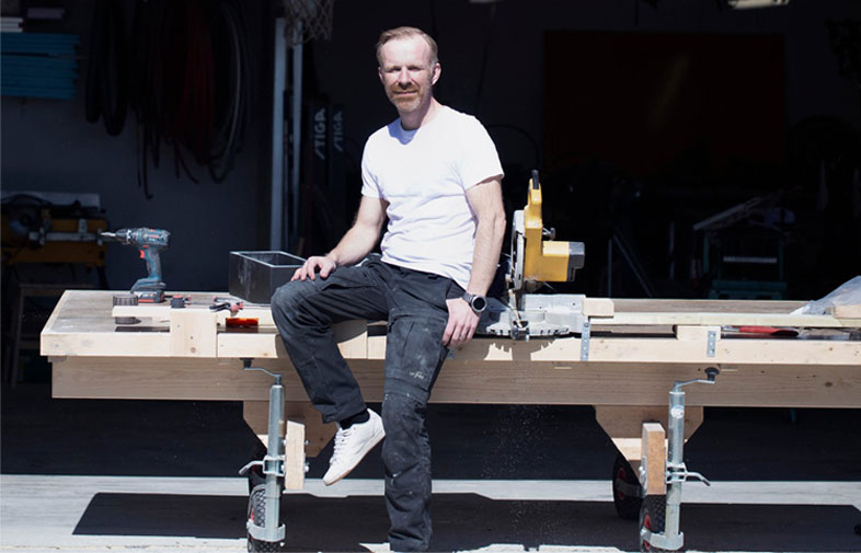 Man i vit tröja och svarta byxor sitter på arbetsbänk med såg och verktyg utanför verkstad