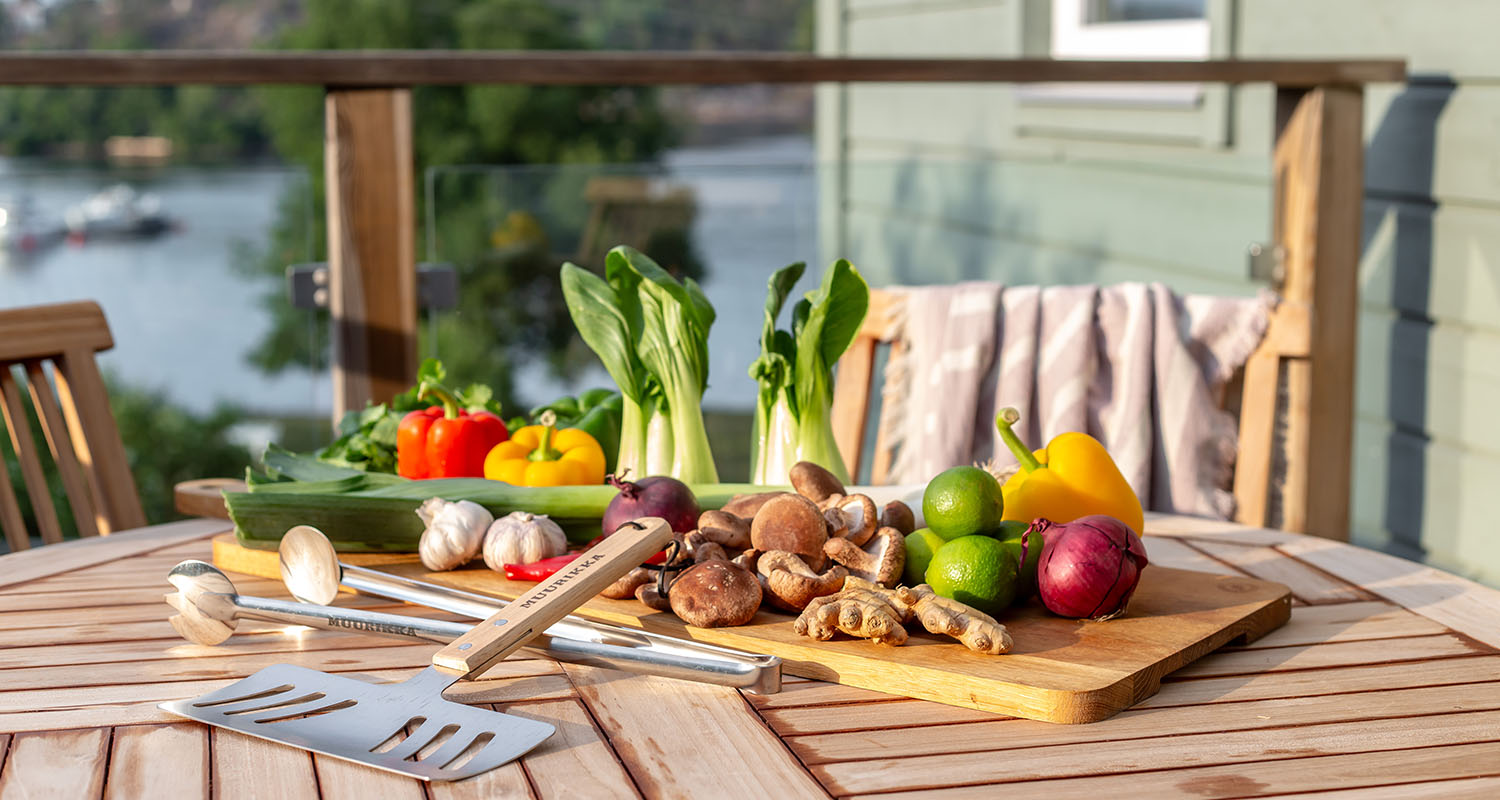 Utebord vid vattnet med skärbräda, paprika, bok choy, lime, svamp och grillredskap.