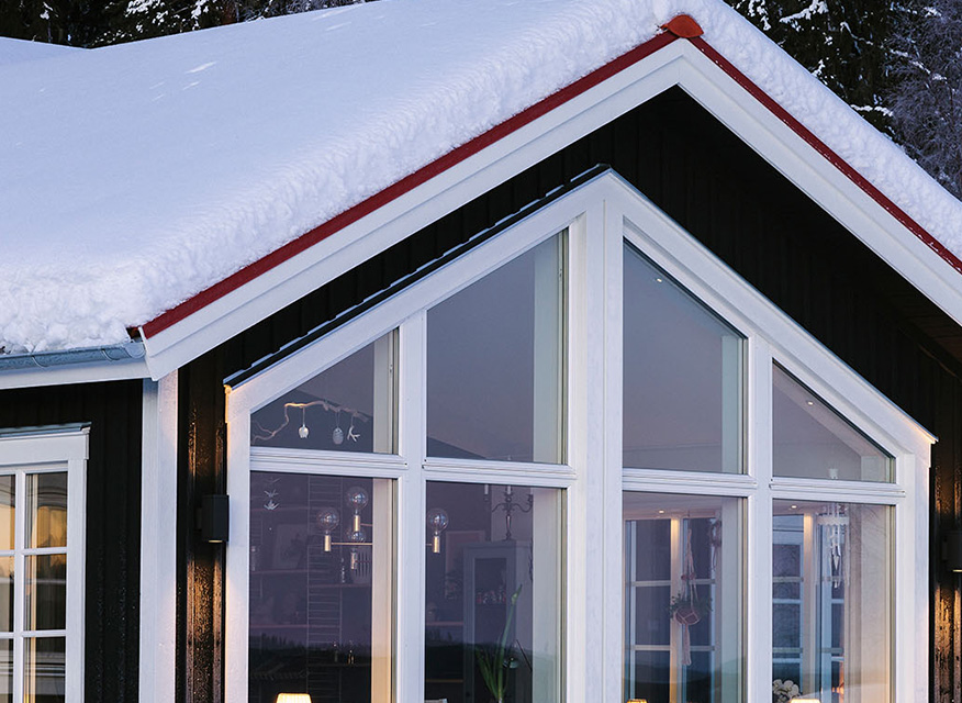 Svart trähus med stora vita fönsterpartier och snötäckt rött tak under vinterkväll.
