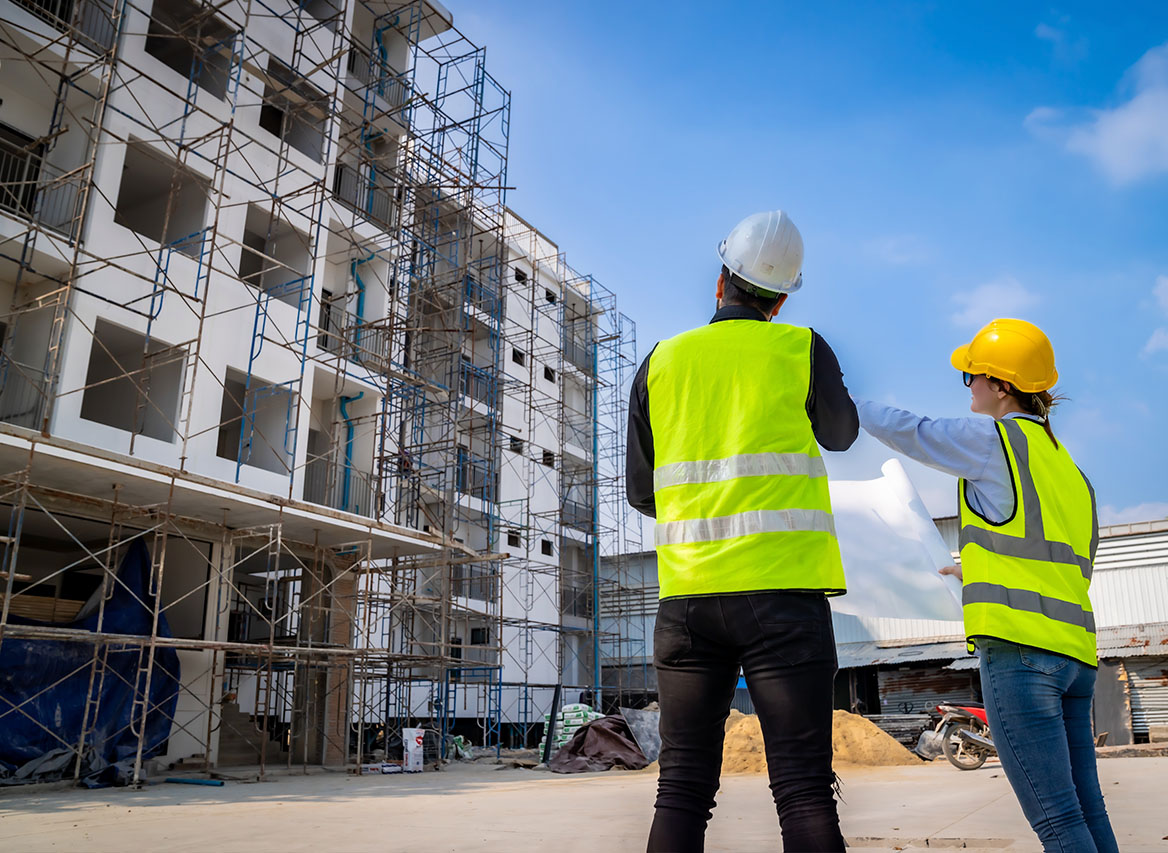 Två byggarbetare i gula västar granskar ritning vid vit byggnad med ställning under blå himmel