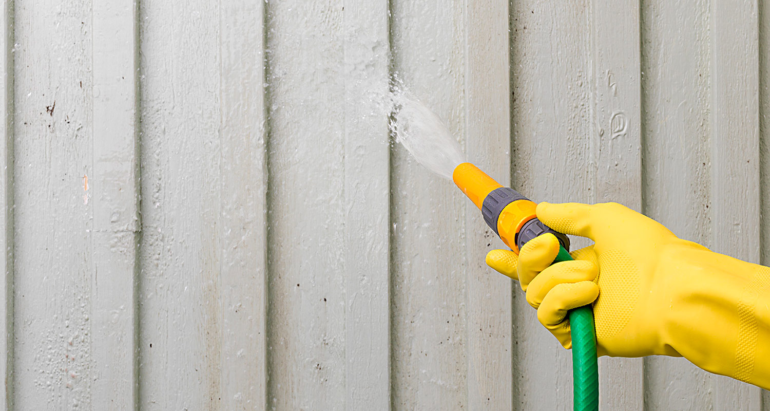 Hand i gul gummihandske som sprutar vatten med grön slang mot grå träpanel