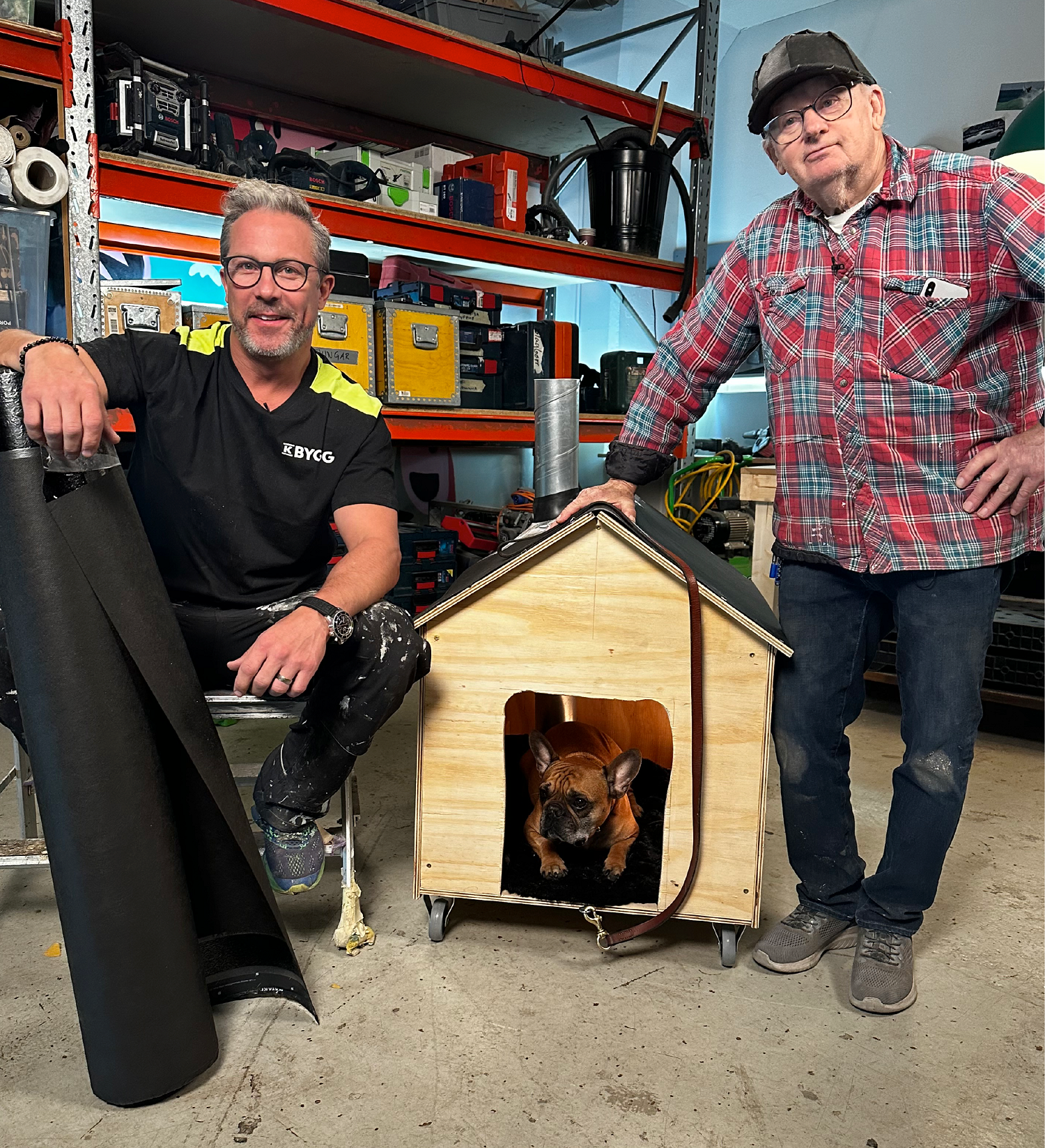 Två män i verkstad med orange hyllor, svart rulle och ljust trähundhus på hjul med fransk bulldogg