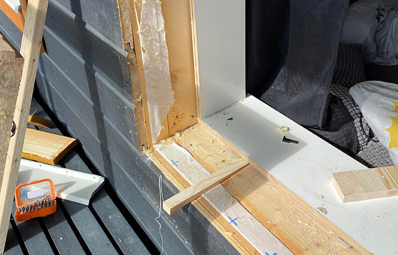 Oinredd träkarm i vit fönsterbänk mot svart fasad, lösa träbitar och orange borrbitsbox.