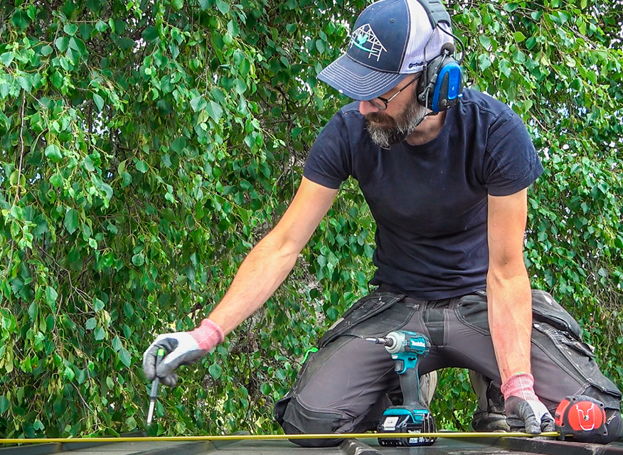 Snickare med hörselskydd mäter på tak med måttband och borrmaskin, bladgrön bakgrund och lila logga
