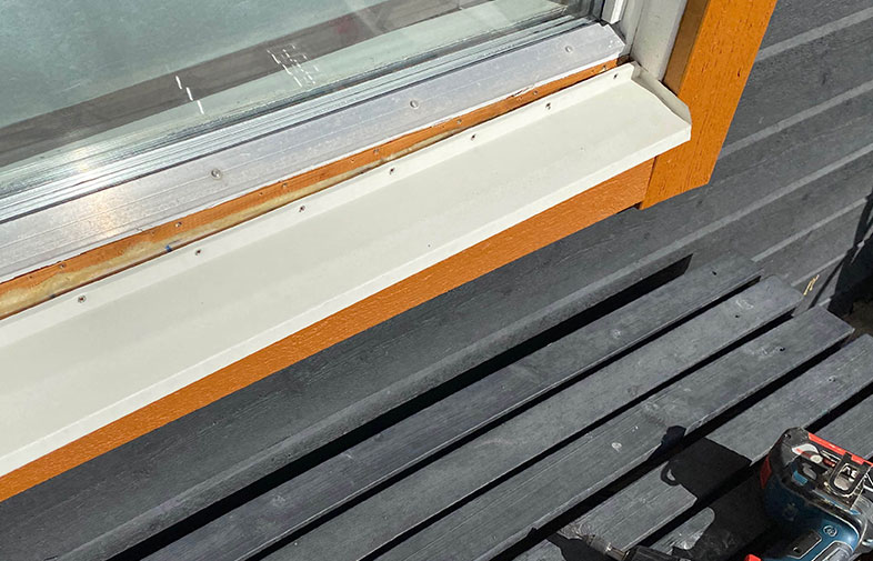 Vitt fönsterbleck monterat under orangebrun karm mot mörkgrå träfasad, borrmaskin syns.