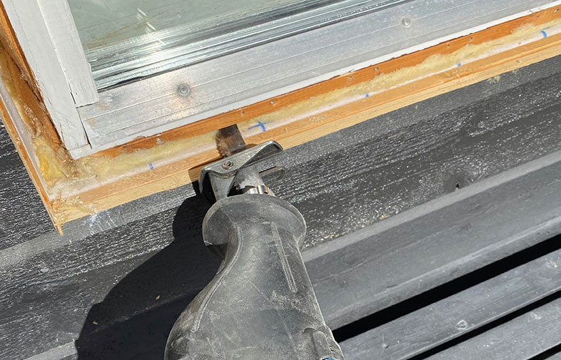 Sticksåg kapar trälist under vit fönsterram mot svart fasadpanel.