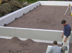 Två män jämnar ut grus i en vit betongform på byggarbetsplats med mätinstrument i bakgrunden.