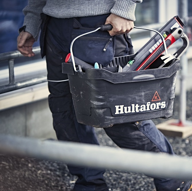 Person i grå tröja bär svart Hultafors-verktygslåda med vattenpass och verktyg