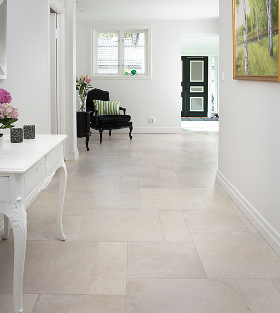 Ljushall med beige klinkergolv, vit hallmöbel, svart fåtölj med grön kudde och blombukett.