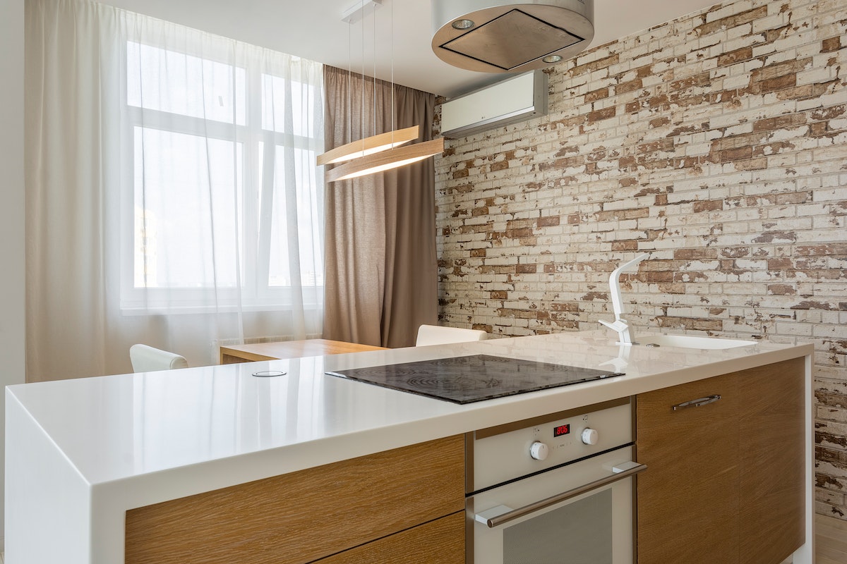 Modernt kök med vit köksö, träskåp, induktionshäll, vitputsad tegelvägg och fönster med gardiner.