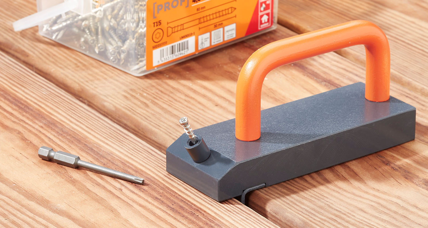 Grå magnetisk skruvhållare med orange handtag och skruv, bits och skruvförpackning på träbord