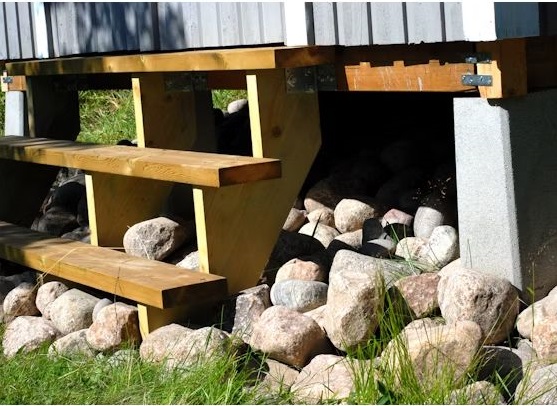 Trätrappa till husgrund vilar på gråa betongpelare, omringad av stora runda stenar och gräs.