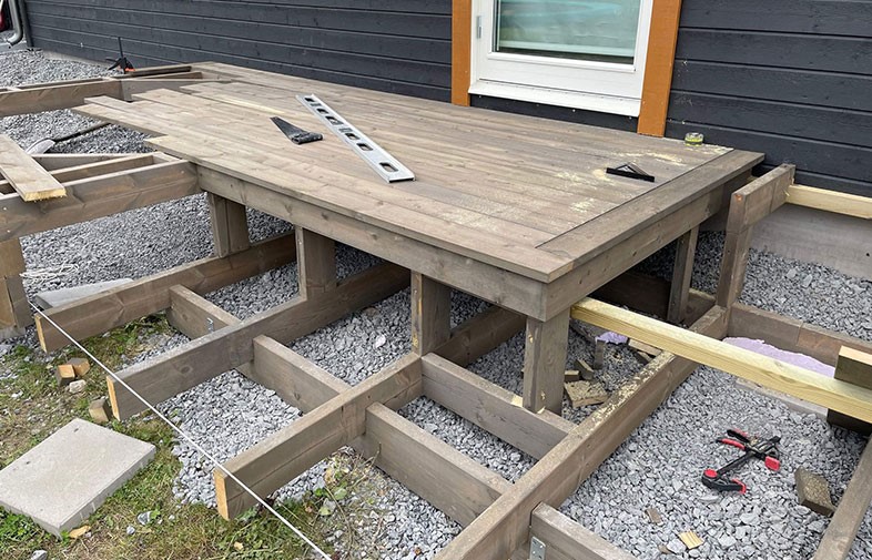 Delvis byggd brun träaltan med reglar på grus och verktyg utanför svart hus med vit dörr