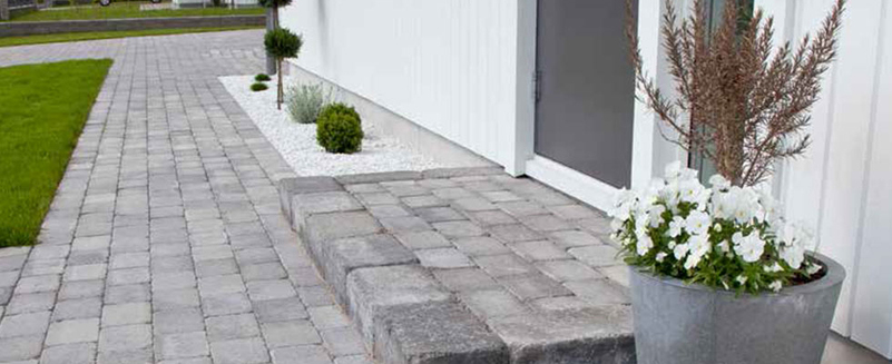 Vit husfasad, grå stenläggning, prydnadsgrus, små gröna buskar och kruka med vita blommor