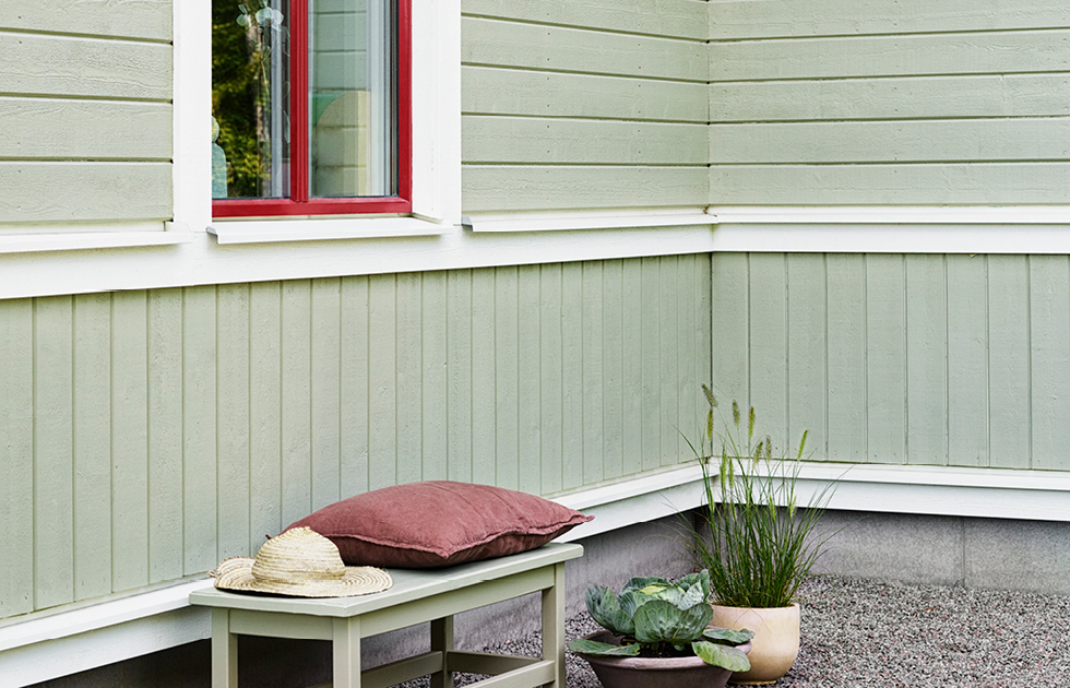 Ljusgrön träfasad med vitt foder och rött fönster, bänk med roströd kudde, stråhatt och krukväxter