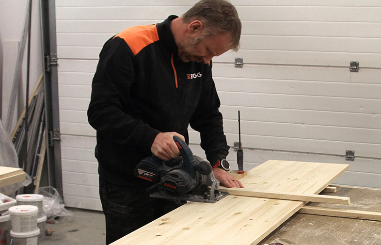 Hantverkare i svart-orange klädsel sågar ljus träskiva med cirkelsåg i verkstad