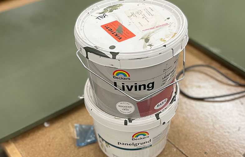 Två vita Beckers-färgburkar (Living, Panelgrund) med gröna färgstänk på trägolv.