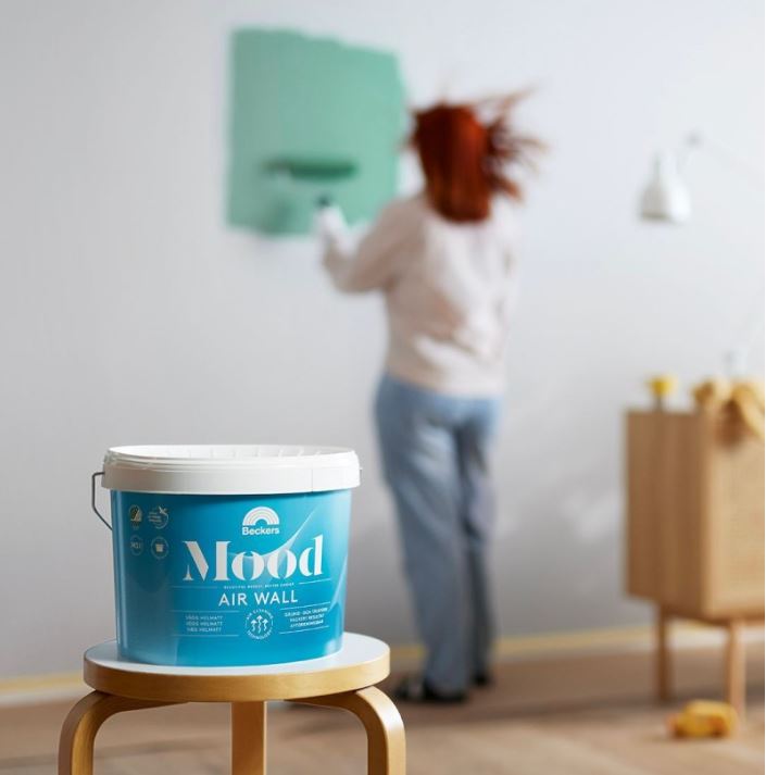 Blå Beckers Mood Air Wall–färgburk på pall i förgrunden, kvinna målar vägg mintgrön.