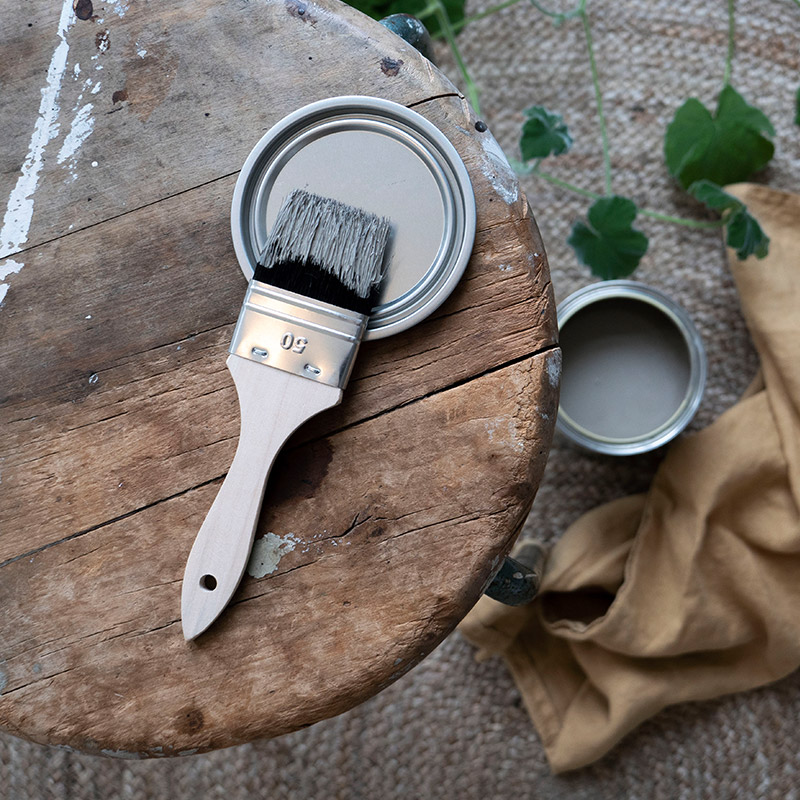 Pensel med grå färg på öppen burk på sliten rund träpall, beige duk och gröna blad.