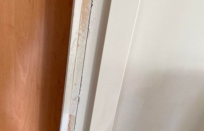 Detaljbild av en sliten vit dörrkarm med flagnande färg intill en orange träddörr.