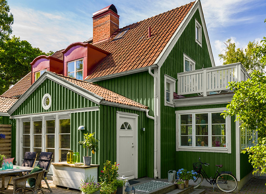 Grönt trähus med vitt fönster och dörr, rött tegeltak, balkong, uteplats med möbler och cykel.