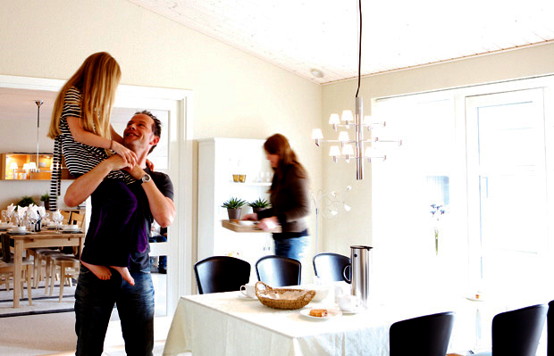 En man håller en flicka i ett ljust rum med vitt matbord, svarta stolar och en kvinna med krukväxter