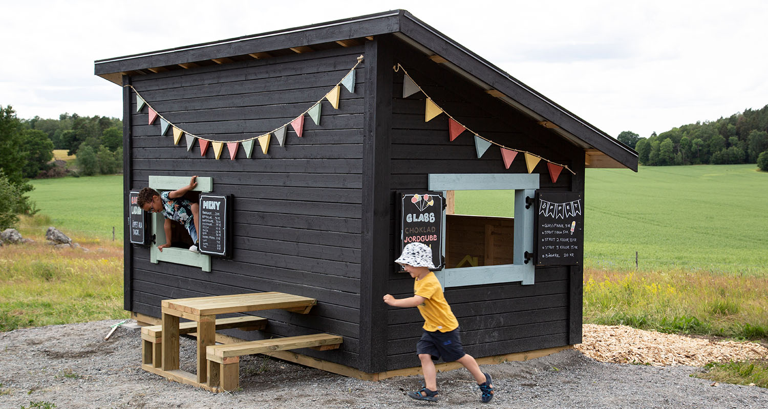 Svart trälekstuga med ljusblå fönster, färgglada vimplar, lekande barn och träbord