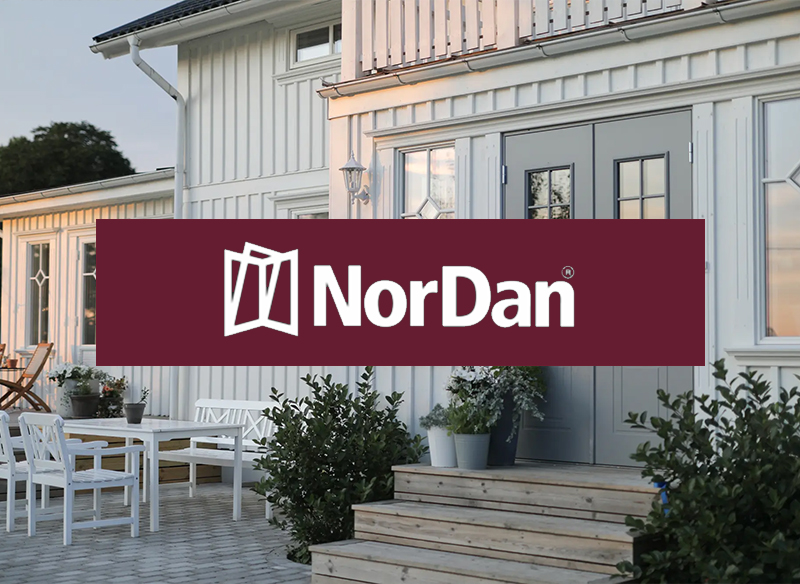 en ljus och klassisk husfasad i träpanel med en grå dubbel ytterdörr. Framför entrén finns en trappa i trä och omgivande gröna buskar, samt en uteplats med vita utemöbler. Ovanpå motivet ligger en vinröd banderoll med NorDan‑logotypen i vitt.