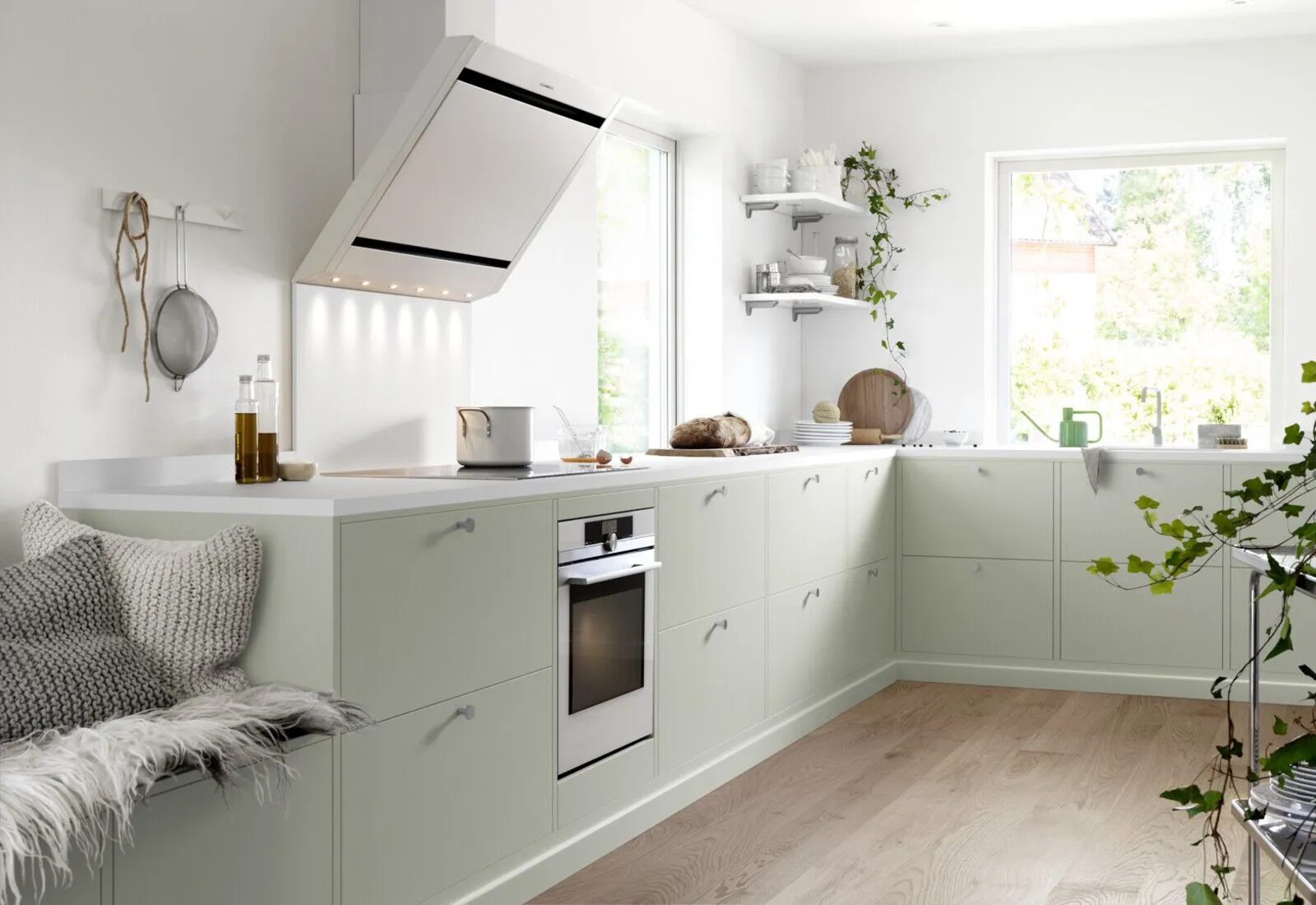 Ljust skandinaviskt kök med mattgröna skåp, vit bänkskiva, rostfri ugn, trädetaljer och växter.