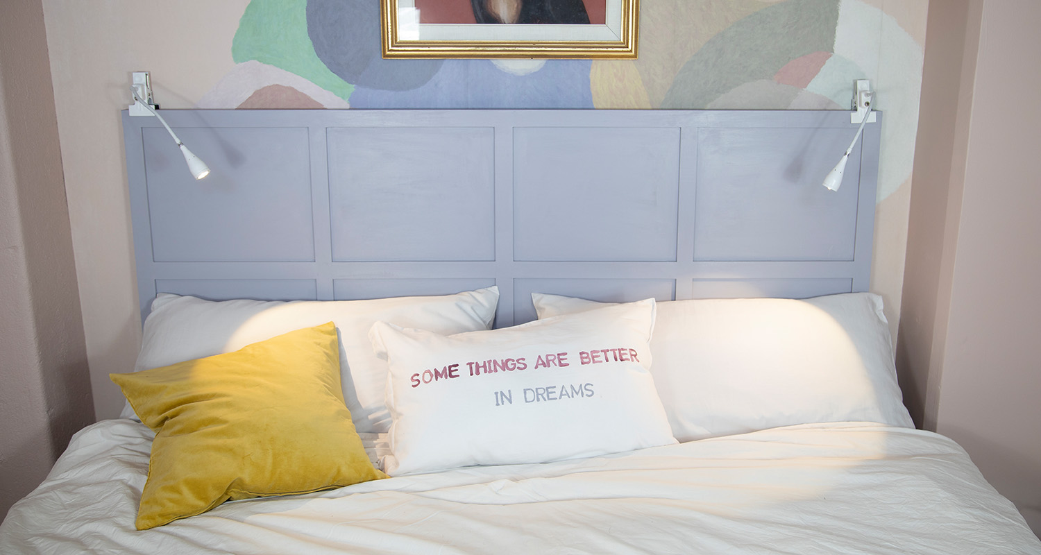Vit bäddad säng med ljuslila gavel, två vägglampor, gul kudde och vit kudde med engelskt budskap