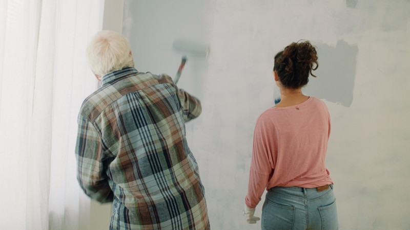 Två personer, äldre man i rutig skjorta och kvinna i rosa tröja, målar en ljusgrå vägg.