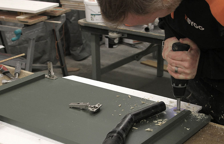 Hantverkare i svart-orange tröja borrar i grön skåpsdörr på arbetsbänk med löst beslag.