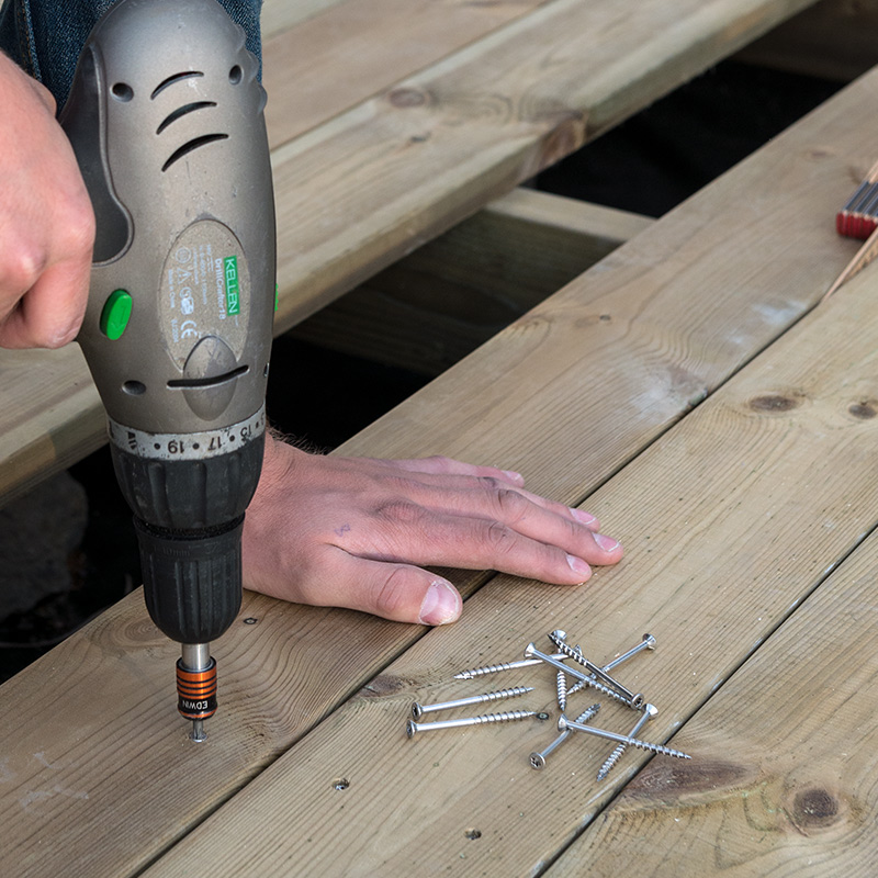 Borrmaskin med gröna detaljer fäster silverljusa skruvar i ljus trallplanka medan hand stöder.