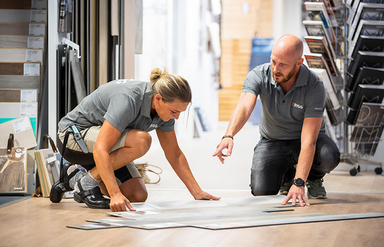 Två personer i grå pikétröjor knästående vid ljusbruna golvprover i butiksmiljö med displayhyllor.