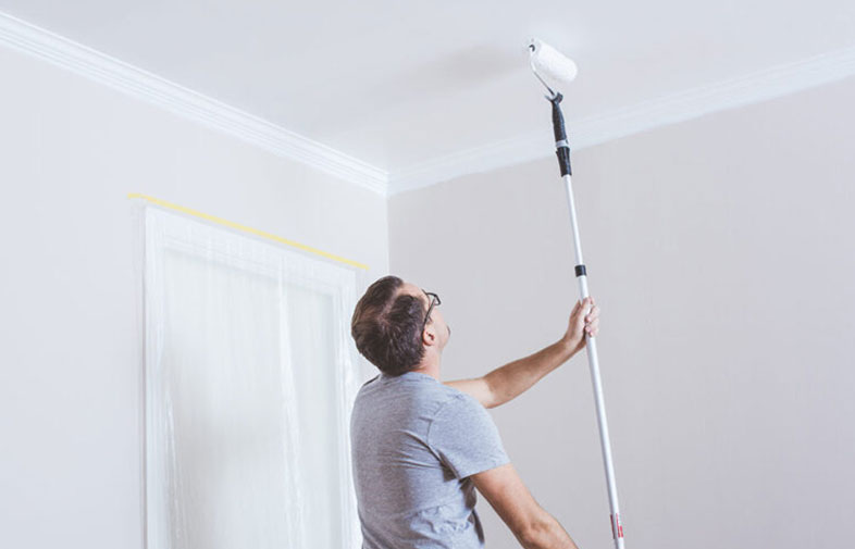 Man i grå t-shirt målar vitt tak med roller på långt skaft i ljusgrått rum.