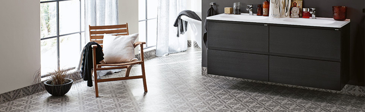 Ljust badrum med stora fönster, grå mönstrad klinkergolv, trästol med vit kudde, svart handfatsskåp.