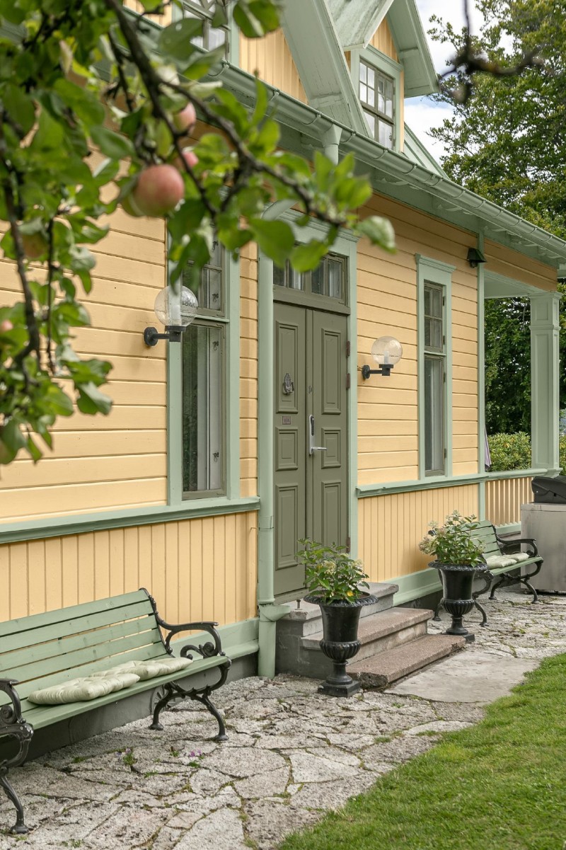 Gul trävilla med grön dörr och fönsterbågar, bänkar och blomkrukor under äppelgren.