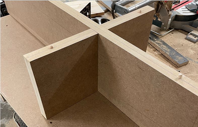 Bruna MDF-skivor korsade i X-form med träplugg på verkstadsbord bland verktyg.