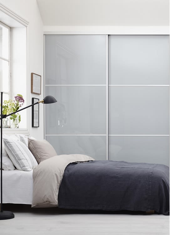 Minimalistiskt sovrum med grå säng, beige påslakan, glas-skjutdörrsgarderob och blommor vid fönstret.