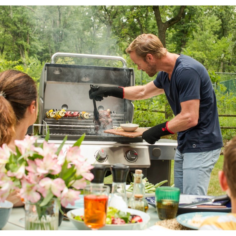 Man i mörk tröja grillar kött på gasolgrill i grön trädgård, dukat bord med rosa blommor.