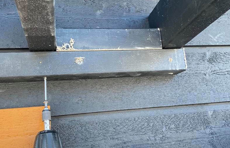 Närbild på svartmålade träbrädor där borrmaskin skruvar i skruv, orange vägg bakom.