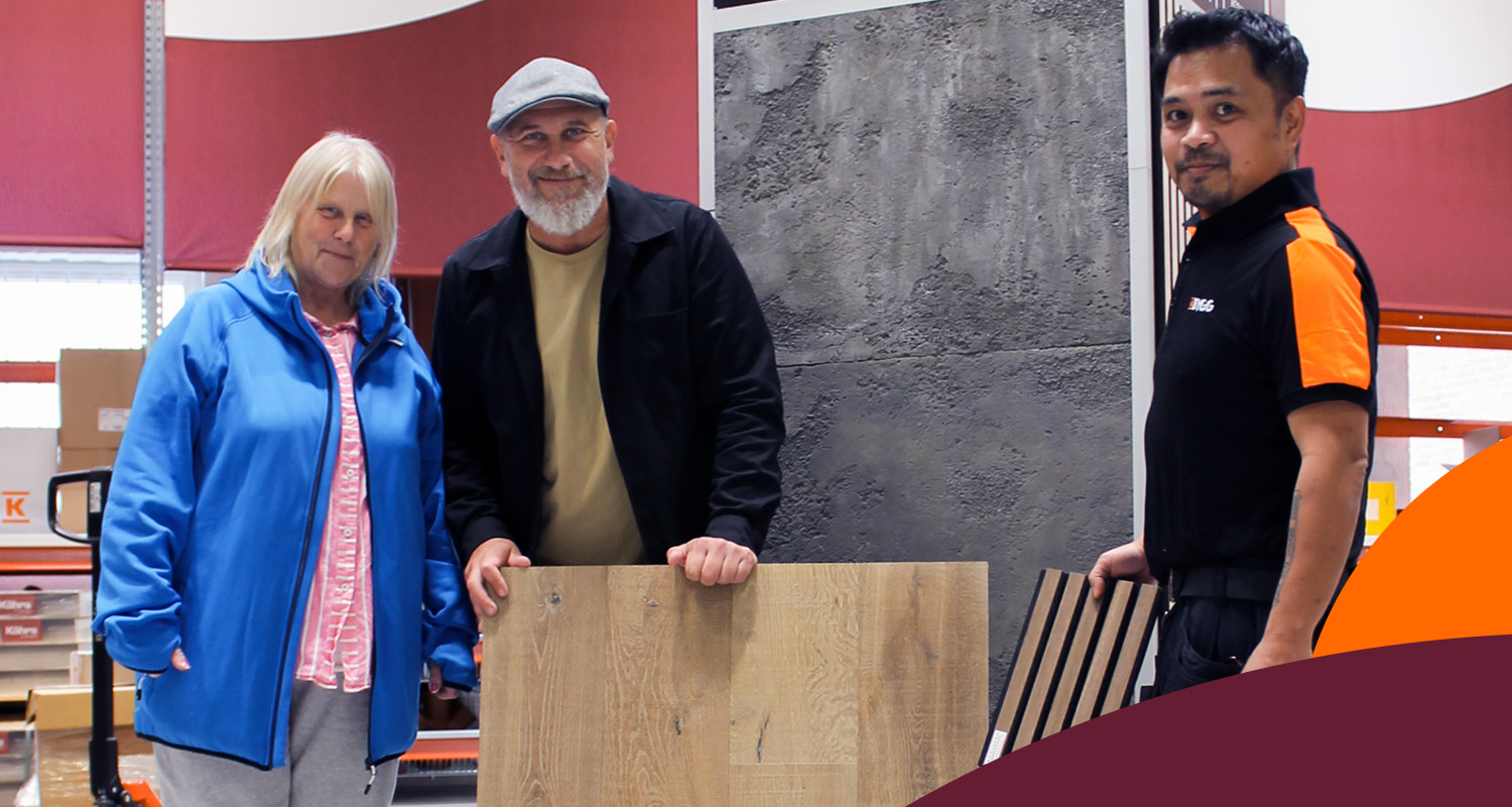 Kundpar och anställd i byggvaruhus framför röd vägg granskar ljus trägolvsskiva