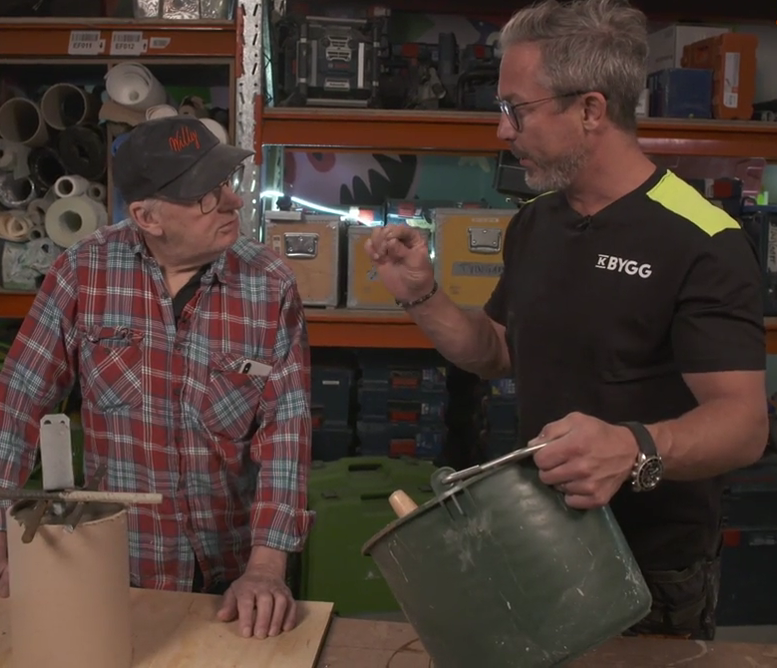 Två män vid bänk i verkstad, en i röd flanell, en i svart tröja med gul axel, med grön hink