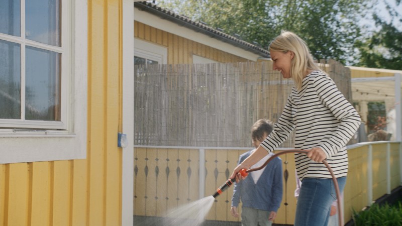 Kvinna i randig tröja vattnar gula husfasaden med slang, barn i bakgrunden.