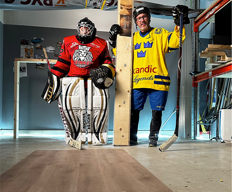 Två ishockeyspelare i röd målvaktsutrustning och gul landslagströja i byggverkstad med träplankor.