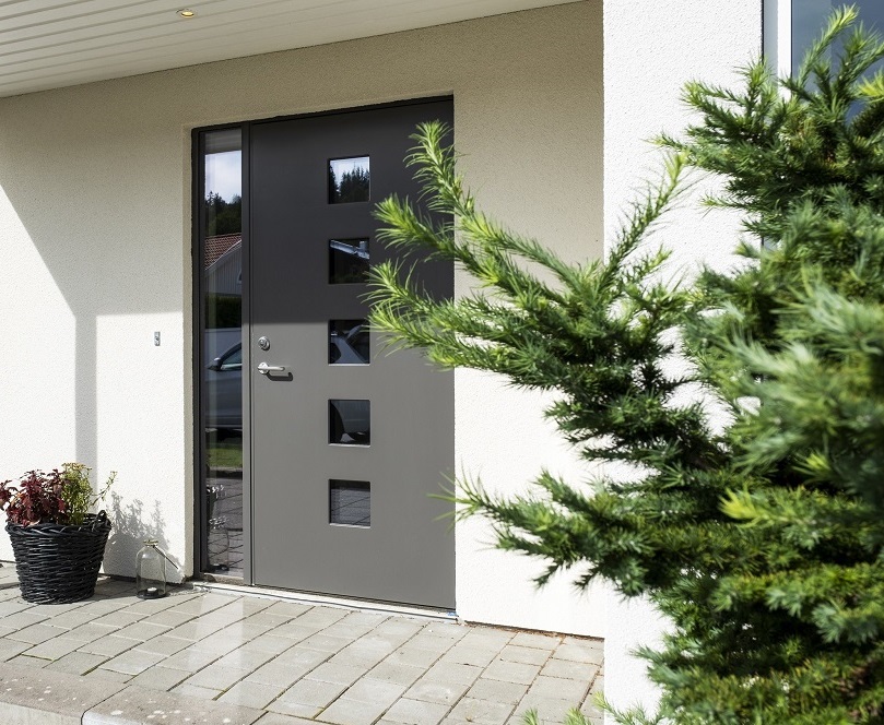 Modern grå ytterdörr med fyra fyrkantiga fönster, vit fasad, grå stenplattor, kruka och grön buske.