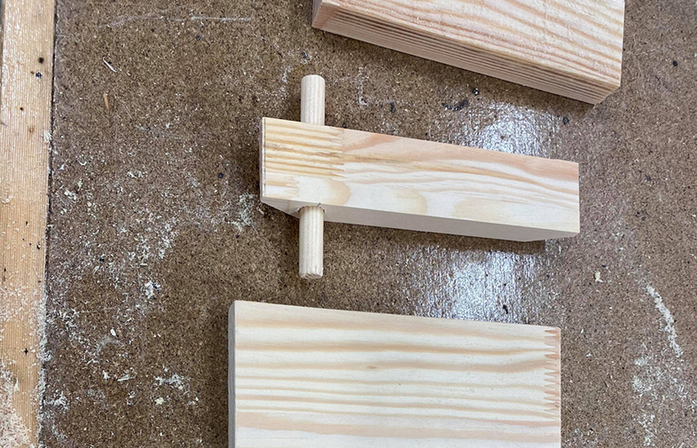 Ljusa träbitar på brun arbetsbänk med sågspån; en bit har ett trästift insatt.