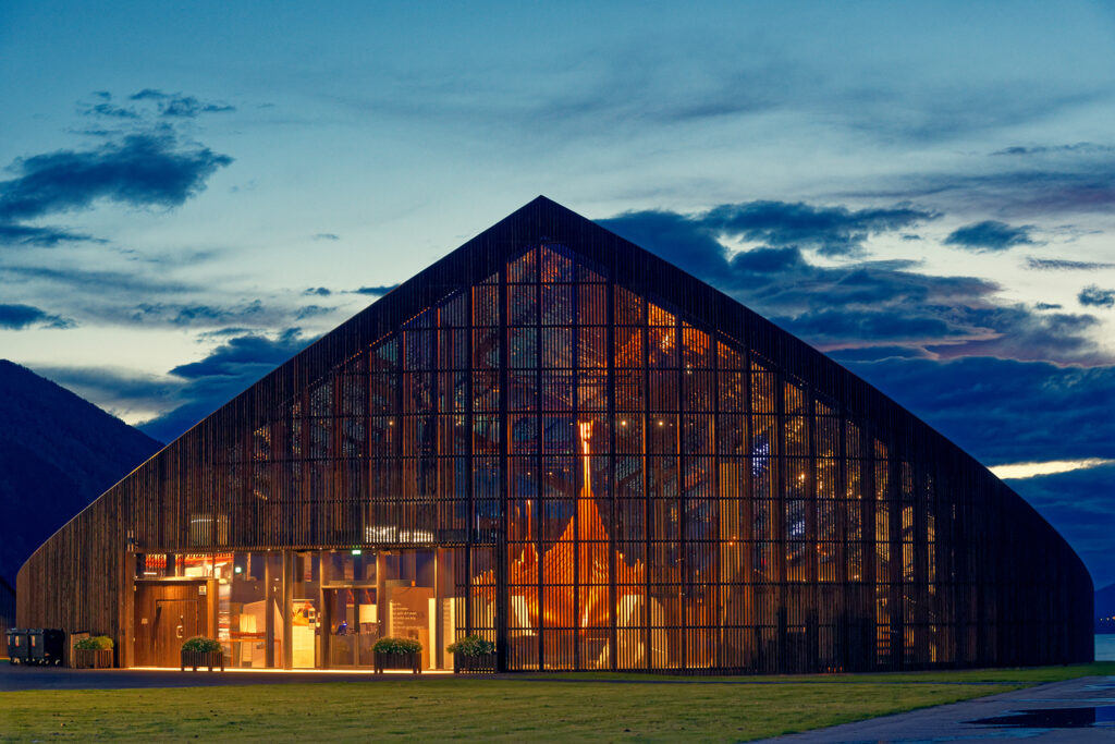 Trekantig glasbyggnad med mörka lameller och varmvitt sken inifrån mot blå skymningshimmel.